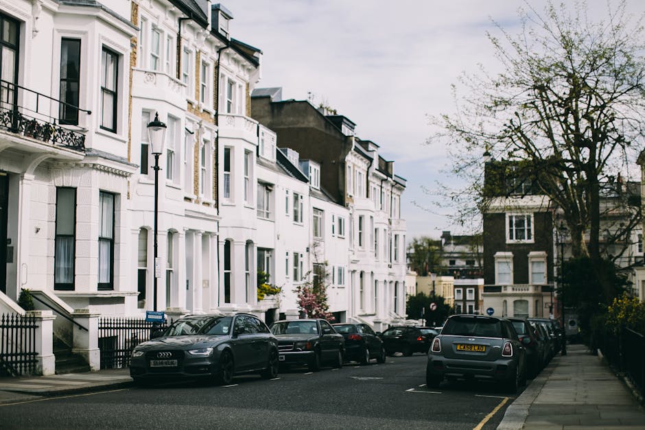 A row of white, multi-storey terraced houses with decorative window frames and small front gardens, situated along a residential street in Gipsy Hill. Parked cars, including an Audi and a Mini Cooper, are lined up along the curb on the left side, with some of the vehicles partially blocking the pavement. A tall, black street lamp is positioned in front of the houses, and a large, leafless tree stands on the right side of the street, casting shadows on the pavement. The scene appears to be overcast, with grey clouds covering the sky. This setting reflects a typical urban neighbourhood suitable for home relocation and furniture transport, often requiring careful planning during the loading process by professionals such as those from Man with Van Gipsy Hill, who manage packing, moving, and vehicle loading within residential areas.