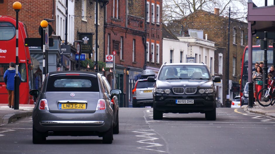 A street scene in Gipsy Hill featuring several parked vehicles, including a silver Fiat 500 with a yellow UK license plate and a black BMW X3 with an blue UK license plate, both parked along the roadside curb. Behind these cars, additional vehicles are visible, including a white van and a silver car, indicating a busy urban environment. The image captures the loading process for a home relocation, with some vehicles possibly used for furniture transport and packing materials, such as cardboard boxes or plastic wrap, although these are not clearly visible. Pedestrians can be seen on the pavement, some near bus stops and street furniture, with buildings lining the background, including shops and residential properties. The scene is well-lit with daylight, emphasizing the urban setting and the logistical considerations involved in moving services offered by Man with Van Gipsy Hill, particularly concerning pre-move permit planning to avoid parking fines in the area.