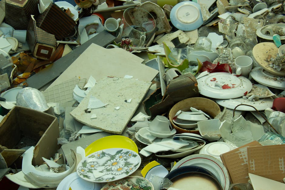A large pile of discarded household items and waste on a pavement, including broken and intact ceramic plates, bowls, cups, and glassware, along with cardboard boxes, plastic packaging, and miscellaneous debris. Some items appear shattered with broken edges and fragments scattered throughout. The background shows a mix of kitchenware and refuse, indicative of waste removal or clearance. This scene reflects the disposal process involved in house removals, such as clearing out household contents before relocation, with a focus on bulky waste management. The image emphasizes the importance of proper waste disposal in house moving services provided by Man with Van Gipsy Hill, supporting efficient and responsible household item removal in SE19.
