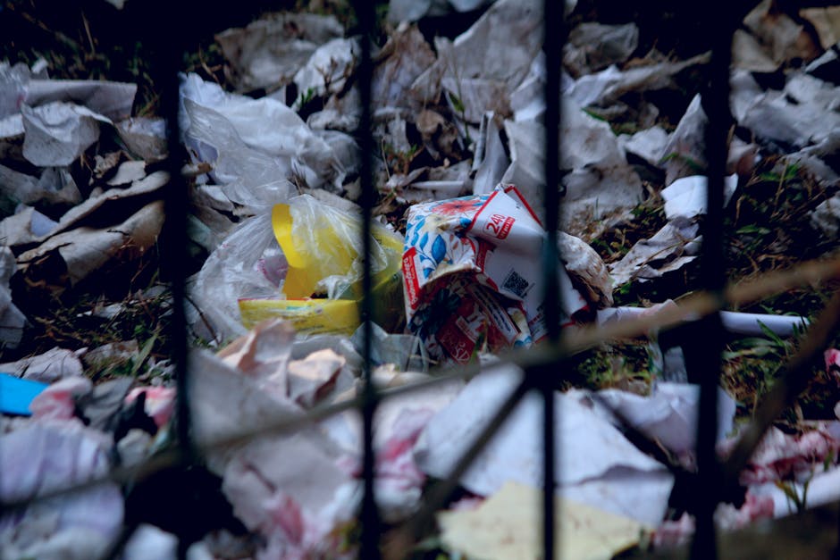 Close-up view of a pile of household waste and packaging materials on the ground behind a black metal fence. The waste includes crushed cardboard boxes, plastic bags, paper, and packaging remnants, situated near the entrance of a property. The scene captures the clutter typical of bulky waste awaiting collection during home relocation or disposal, with some items partially obscured by the fence bars. The surrounding environment suggests an outdoor area on pavement or a garden edge. Man with Van Gipsy Hill offers house removals and waste clearance services, providing professional assistance with packing, transporting, and managing bulky household waste as part of moving logistics or disposal processes in SE19 area, such as at GIPSY HILL.