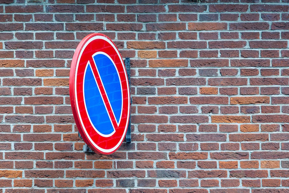 A round, red and blue no stopping or parking sign mounted on a black metal bracket is affixed to a red brick wall exterior. The sign features a blue circle with a red border and a red diagonal line cr