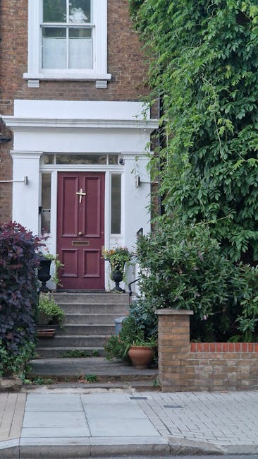 The image shows the front entrance of a Victorian Gipsy Hill home, featuring a maroon wooden front door with a brass letterbox and a gold handle, accessible by a small set of concrete steps with a metal handrail on the left side. The door is flanked by white decorative moulding and framed by a white facade, with a small window above it. To the right of the entrance, there is dense green foliage, including a large leafy tree, partially obscuring the brick wall and garden area. Potted plants and a small shrub are positioned on either side of the steps, enhancing the residential appearance. The pavement leading to the house is paved with grey concrete slabs, and the property’s brick wall extends along the right edge. This setting illustrates a typical home exterior in an urban area, relevant to house removals and the logistical considerations involved in packing and transporting furniture, especially in homes with narrow staircases, as referenced in the page title.