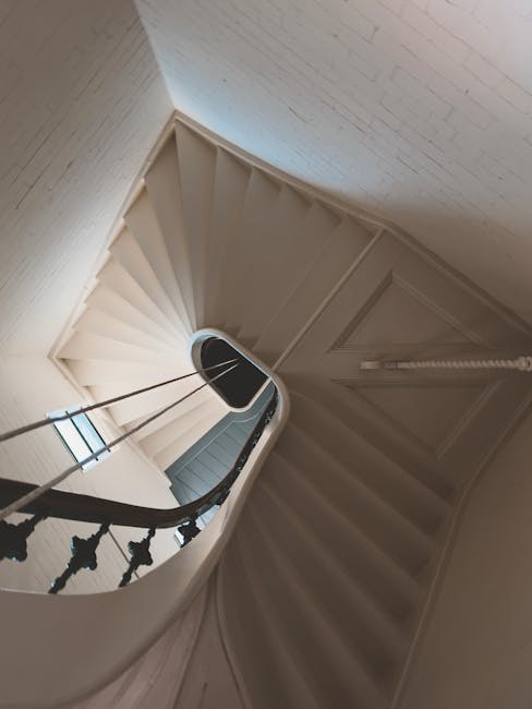 A spiral staircase inside a property with white painted walls and wooden handrails supported by metal balusters. The staircase curves downward from an upper floor, with a small landing visible at the top. Natural light enters through a window at the bottom of the staircase, illuminating the area. The walls are finished with textured wallpaper, and a white-painted wooden door is slightly ajar at the top of the stairs. The image captures a clean, well-maintained interior space, with no furniture or belongings visible, illustrating a typical residential staircase suitable for a house relocating service like Man with Van Gipsy Hill, focusing on the navigation and space considerations when moving furniture or boxes between floors.