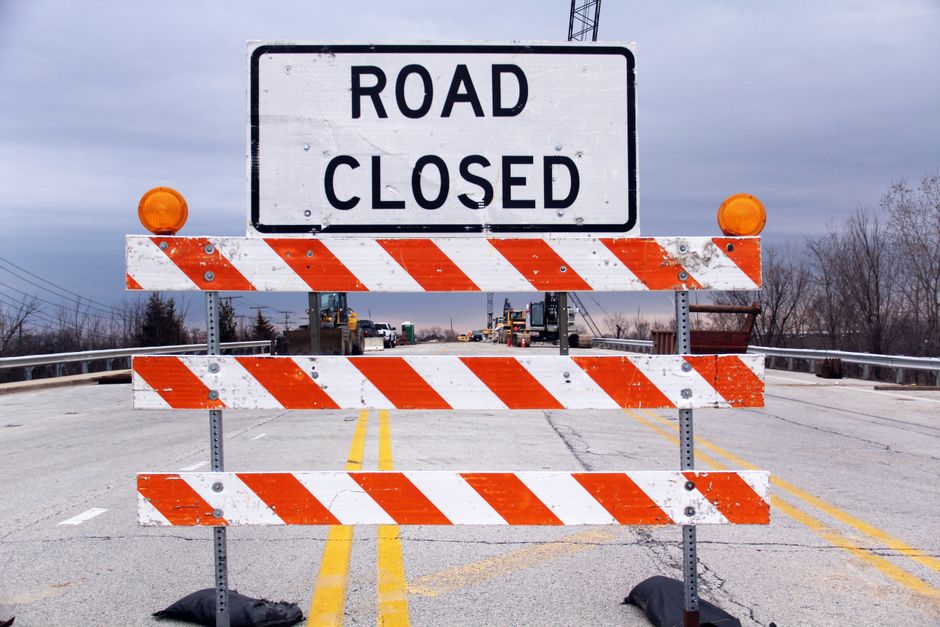 A road closed sign with two orange warning lights mounted on a metal frame, positioned across a two-lane road designed for a home relocation or furniture transport. Behind the sign, the road is blocked with a striped orange and white barrier, and the pavement has yellow lane markings. In the distance, construction vehicles and equipment are visible, suggesting ongoing road work or a permit process for moving services. Bare trees and overcast sky can be seen on the horizon, indicating outdoor conditions suitable for loading or unloading moving boxes, furniture, and appliances. The scene captures the logistical considerations involved in traffic management during residential removals, highlighting the importance of permits and planning for house relocations. Man with Van Gipsy Hill occasionally provides such services, including navigating road closures for smooth and efficient moves.