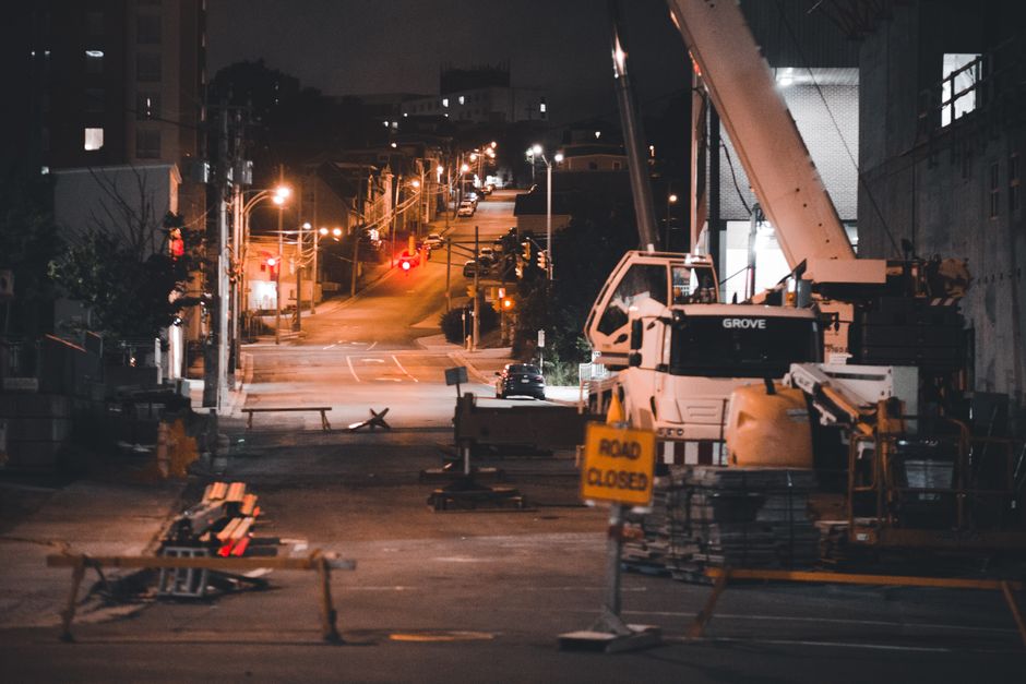 A nighttime view of a street undergoing construction, with a large white mobile crane positioned on the right side of the image on a paved section of road, surrounded by orange and white barricades and a yellow 'Road Closed' sign. To the left, stacks of construction materials such as metal beams or pipes are placed on stands, and some are wrapped in plastic or fabric. In the background, the street slopes uphill with streetlights casting orange illumination on parked cars and buildings lining both sides. The scene captures the process of road closure and construction activity, relevant to local house removals and moving logistics in the Gipsy Hill area, as handled by Man with Van Gipsy Hill.