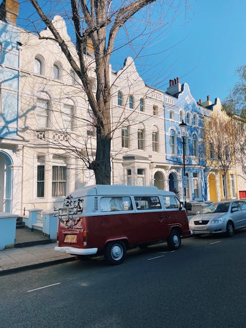 A vintage light blue Volkswagen Type 2 van is parked on a residential street, with a roof rack mounted on top. The van's front windshield reflects nearby trees with yellow and green foliage, and it ha