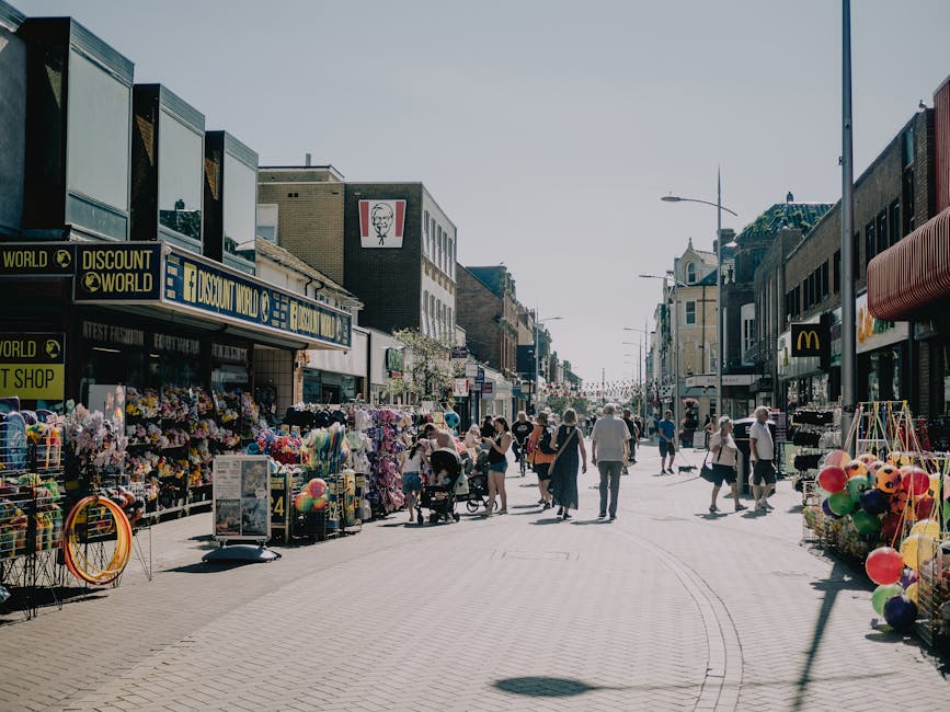 A busy street scene during daylight hours in Gipsy Hill with storefronts including a discount shop, a fast-food restaurant, and various retail outlets visible. The pavement is populated with pedestrians, some walking, others pushing strollers or carrying shopping bags, and several individuals are engaged in conversations. On the left side, a display of outdoor market stalls holds colorful toys, balloons, and household items arranged in groups, with some items on stands and others in baskets. To the right, similar street vendors display balloons and other merchandise, with a McDonald's sign visible on a building further down the street. The environment appears vibrant, with clear weather and natural lighting casting soft shadows on the ground. This scene illustrates typical urban activity associated with residential area relocations or moving logistics, where transport or packing processes might involve accessing nearby shops or street-view premises nearby [manwithvangipsyhill.co.uk], reflecting a typical move or delivery setting related to house removals in Gipsy Hill.