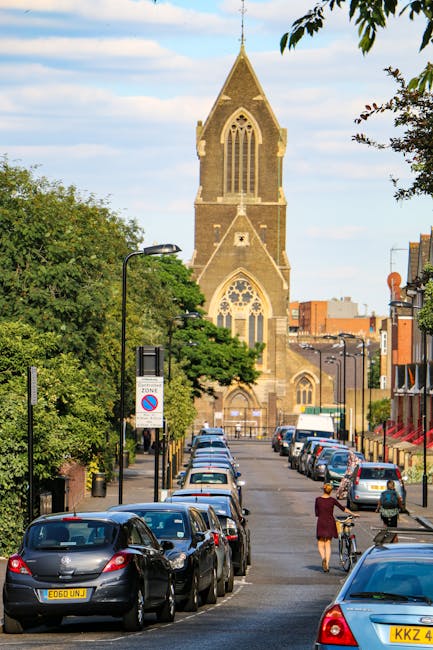 A street scene in Gipsy Hill featuring parked cars lining both sides of the road in the foreground, with a woman walking her bicycle and another pedestrian nearby. On the left side, there are trees and a street lamp, while on the right, there are buildings with shopfronts. In the background, a historic stone church with a tall, pointed spire and large arched windows dominates the scene, set against a blue sky with scattered clouds. The image depicts a typical residential and commercial area relevant to house removals and moving services, with clear visibility of vehicles, street furniture, and architectural details. Occasionally, Man with Van Gipsy Hill may assist with home relocation and furniture transport, ensuring the smooth logistics of packing and moving in the area.