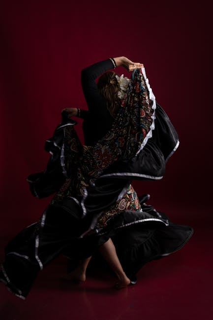 A person with long hair is performing a dynamic dance move against a dark red background. They are wearing a black long-sleeved top and loose black pants. Their movements involve a swirling motion with a large piece of fabric or clothing, which has intricate, colorful patterns and white edging, creating a flowing, layered effect. The fabric appears to be lifted and twisted in mid-air, emphasizing the motion and energy of the dance. The person's feet are bare, and their body is balanced on one foot with the other slightly raised, capturing a moment of agility and grace. The scene is well-lit, highlighting the textures of the fabric and the contours of the dancer’s form. This image visualizes expressive movement and fabric dynamics, reminiscent of home relocation or packing and moving processes involving fabric coverings or protective clothing used during furniture transport, which ties subtly into the themes of moving services by Man with Van Gipsy Hill.