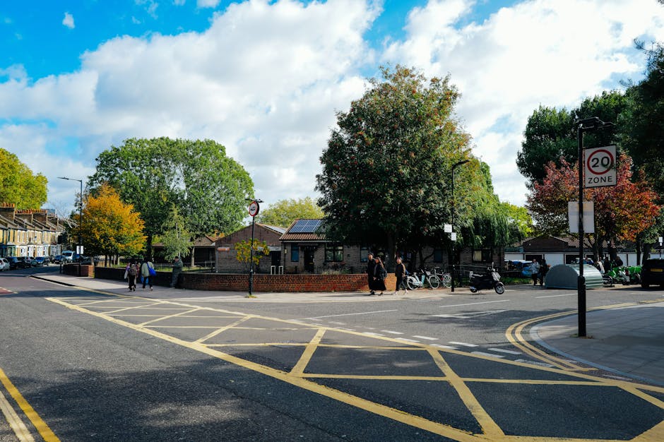 A street scene in Gipsy Hill featuring parked cars lining both sides of the road in the foreground, with a woman walking her bicycle and another pedestrian nearby. On the left side, there are trees and a street lamp, while on the right, there are buildings with shopfronts. In the background, a historic stone church with a tall, pointed spire and large arched windows dominates the scene, set against a blue sky with scattered clouds. The image depicts a typical residential and commercial area relevant to house removals and moving services, with clear visibility of vehicles, street furniture, and architectural details. Occasionally, Man with Van Gipsy Hill may assist with home relocation and furniture transport, ensuring the smooth logistics of packing and moving in the area.