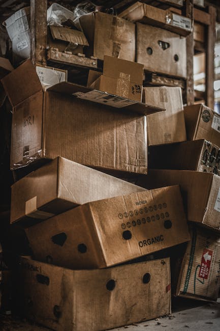 A collection of cardboard boxes of various sizes stacked on top of each other inside a storage area, with some boxes showing printed labels and symbols. The boxes are made of brown corrugated cardboard, with some sealed with packing tape and others open or partially collapsed. They are placed on the floor and on wooden shelves, indicating an organized packing or storage process related to house removals. The environment appears to be illuminated by natural or ambient light, with a focus on the packed items prepared for a home relocation or furniture transport. The scene reflects the packing and moving logistics handled by Man with Van Gipsy Hill, highlighting the careful handling and organization involved in household removals.