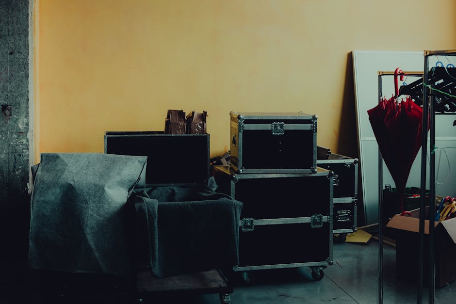 Inside a household room during a home relocation process on Coldharbour Lane, several black storage cases with metal reinforced edges are stacked on the floor near a yellow wall, indicating the packing and loading phase. Nearby, a piece of furniture covered with grey protective fabric and a foam padding is positioned adjacent to the cases, suggesting preparation for moving. To the right, a metal clothing rack holds hanging red umbrellas and other items, with a white cabinet or mirror partially visible behind it. Items such as cardboard boxes, yellow packing paper, and various tools are present on the floor and in a nearby open box, reflecting the packing materials used for a furniture transport or house removals service. The environment appears to be within a property, with natural lighting highlighting the objects, illustrating the process of loading and packing bulky household items for a professional move, as carried out by Man with Van Gipsy Hill.