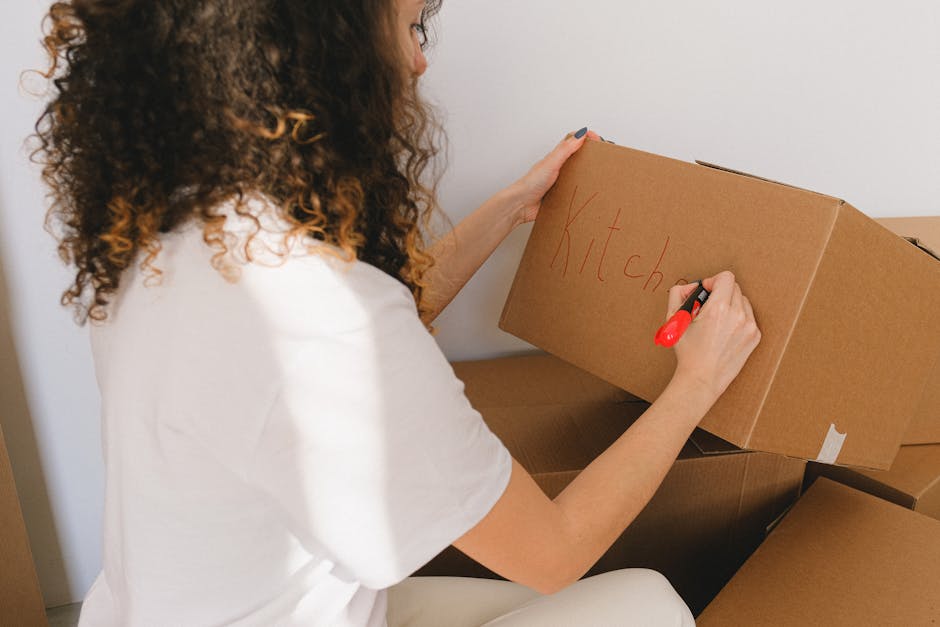 A collection of cardboard boxes of various sizes stacked on top of each other inside a storage area, with some boxes showing printed labels and symbols. The boxes are made of brown corrugated cardboard, with some sealed with packing tape and others open or partially collapsed. They are placed on the floor and on wooden shelves, indicating an organized packing or storage process related to house removals. The environment appears to be illuminated by natural or ambient light, with a focus on the packed items prepared for a home relocation or furniture transport. The scene reflects the packing and moving logistics handled by Man with Van Gipsy Hill, highlighting the careful handling and organization involved in household removals.