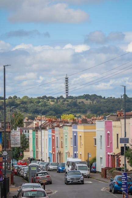 A row of colorful Victorian-style terraced houses in Gipsy Hill, each painted in pastel shades including yellow, pink, blue, green, and purple, lining a narrow urban street filled with parked cars and a few moving vehicles, with some houses featuring small front gardens and brick walls. In the background, a hill with lush greenery and trees is visible, topped by a large communication tower under a partly cloudy sky with patches of blue, illustrating a typical residential neighbourhood suitable for home relocation and furniture transport services such as those offered by Man with Van Gipsy Hill.