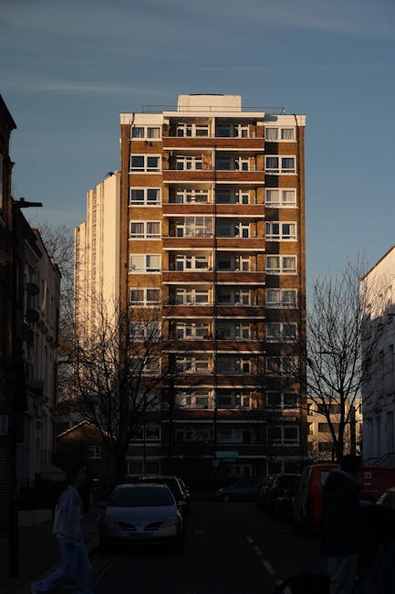 Photograph of a multi-storey residential building with a brown brick and concrete facade, featuring multiple balconies with glass railings and large windows, set against a blue sky with some clouds. The building is situated on a street with parked cars along the pavement, and leafless trees line the road indicating a colder season. In the foreground, there are a few pedestrians walking, and the street includes a sidewalk and street lamps. This urban scene illustrates the typical environment for home relocation or furniture transport, with a focus on an apartment building that may require professional removals services, such as those offered by Man with Van Gipsy Hill, to facilitate packing, loading, and moving belongings in and out of flats near Norwood Road and GIPSY HILL.