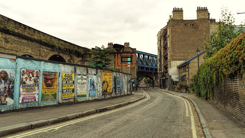 A row of colorful Victorian-style terraced houses in Gipsy Hill, each painted in pastel shades including yellow, pink, blue, green, and purple, lining a narrow urban street filled with parked cars and a few moving vehicles, with some houses featuring small front gardens and brick walls. In the background, a hill with lush greenery and trees is visible, topped by a large communication tower under a partly cloudy sky with patches of blue, illustrating a typical residential neighbourhood suitable for home relocation and furniture transport services such as those offered by Man with Van Gipsy Hill.