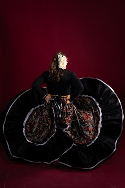 A woman dressed in traditional flamenco attire stands with her back facing the camera against a deep red background. She is wearing a black long-sleeved top and a voluminous, decorated skirt with intricate red and gold floral patterns. Her dark hair is styled in loose waves, adorned with a large white flower on the right side of her head. She holds the sides of her skirt, lifting it slightly to reveal the richly patterned fabric, which appears to be made of satin or silk with a glossy finish. The skirt's structure is supported by layers of fabric, creating a dramatic, full silhouette, reminiscent of traditional Spanish dance costumes. Her pose and the styling suggest a performance or cultural dance context, emphasizing movement and elegance in a studio setting. This image is relevant for showcasing traditional dance costumes, event performances, or cultural celebrations related to Spain.