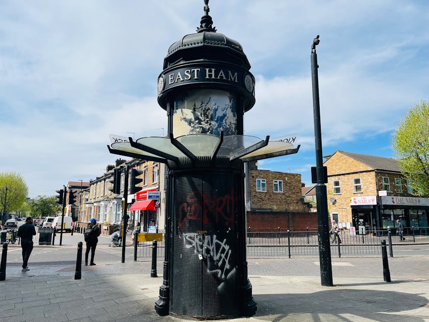 A tall, black, vintage-style informational kiosk with a rounded top and a domed roof displaying the words 'East Ham' on a busy street corner. The kiosk features a glass display panel with posters or advertisements, surrounded by graffiti and stickers. It is positioned on a paved sidewalk with several people walking nearby, some holding bags or umbrellas. The background shows residential and commercial buildings, including a nail salon and other small shops, with trees and a blue sky with some clouds overhead. Traffic lights and street signage are visible, indicating an urban environment. This setting illustrates a typical town centre scene with pedestrian activity and street furniture, relevant to home relocation and moving logistics in city areas, with the kiosk serving as a recognizable landmark in the street scene.